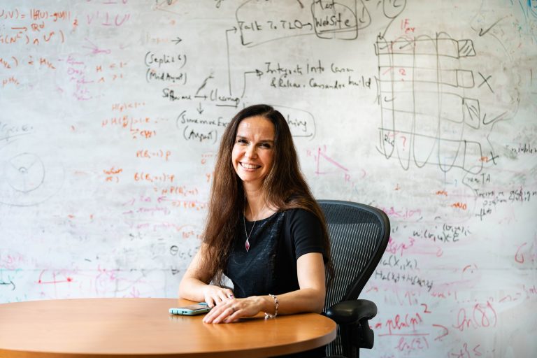 MIT Professor Muriel Médard sitting at a table in front of a diagram-covered whiteboard (Optimum)