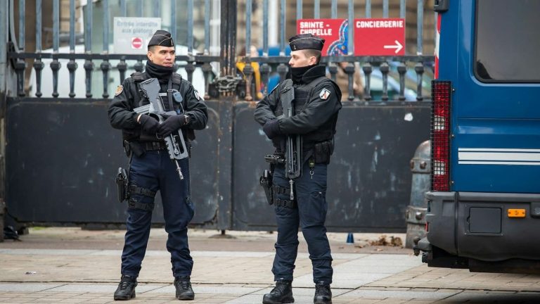 Two men in uniform carrying submachine guns and other arms stand outside an iron gate.