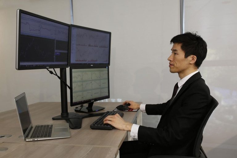 A besuited trader monitors three desktop screens and a laptop. (harryloya/Pixabay)