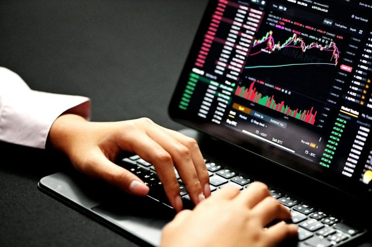 A pair of hands resting on a keyboard with an iPad showing graphs and price quotes. (Kanchanara/Unsplash)